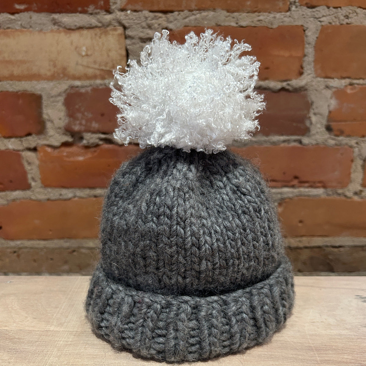A dark gray knitted beanie hat with a ribbed cuff and a fluffy white pom-pom, displayed on a light wooden surface in front of a brick wall background.
