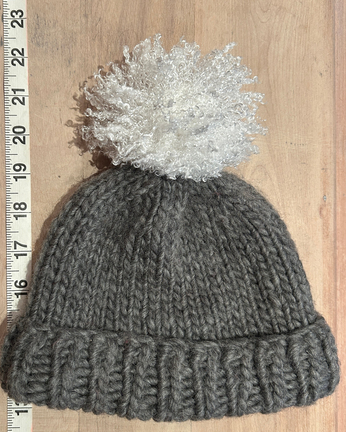 A gray ribbed knit beanie with a fluffy white pom-pom, laid flat on a wooden surface next to a measuring tape ruler.