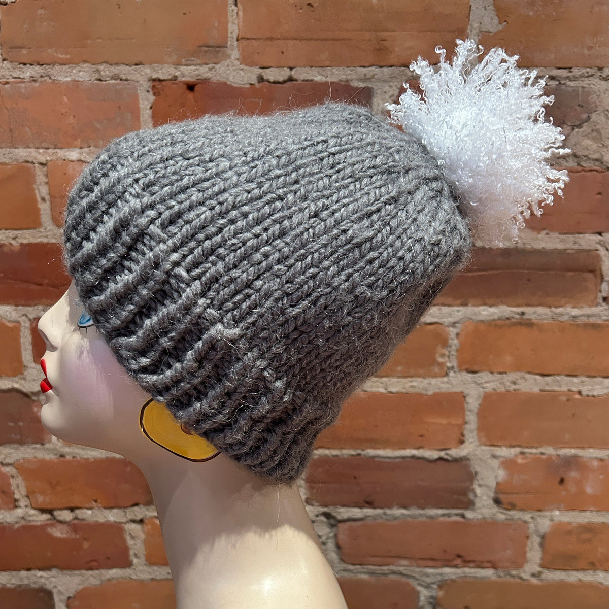 A gray ribbed knit beanie with a fluffy white pom-pom, displayed on a mannequin head against a red brick wall background.