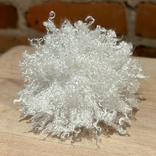 A fluffy, curly white decorative pom-pom or fiber ball placed on a light wood surface with a blurred brick wall background.