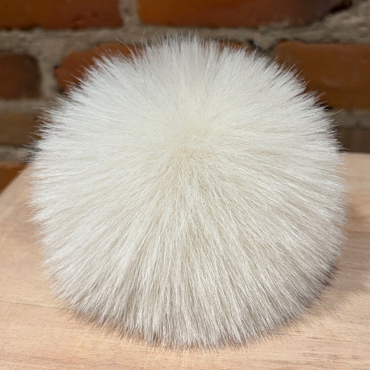 A fluffy ultra light beige faux fur pom-pom ball resting on a light wood surface with a blurred brick wall background.