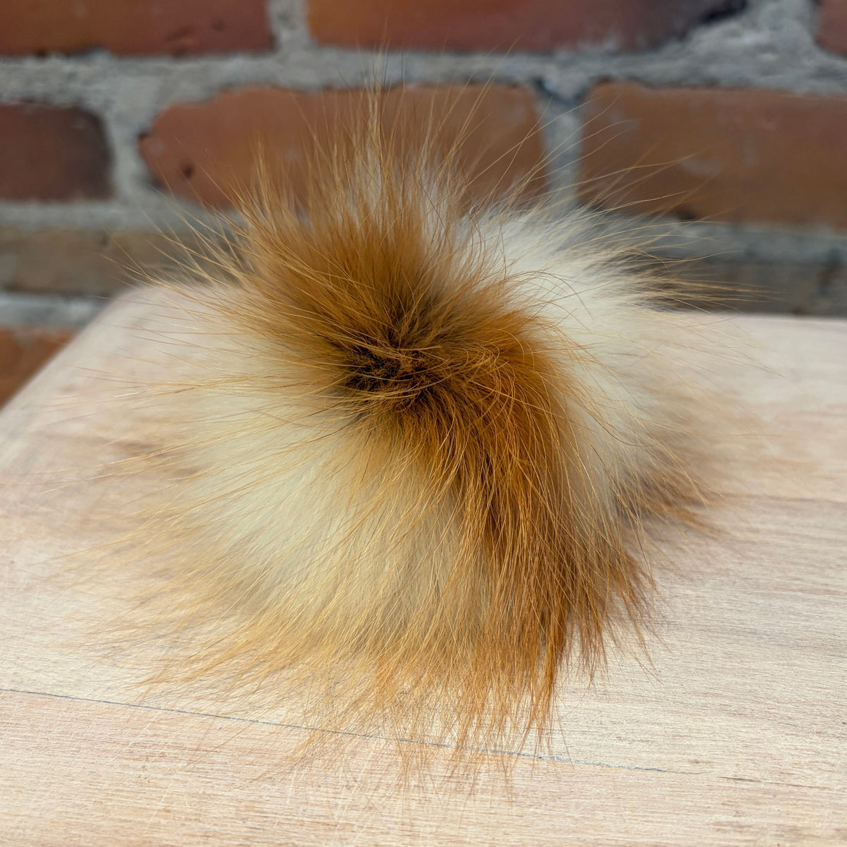 A fluffy fox fur pom-pom accessory with a mix of cream, light brown, and reddish-brown tones, resting on a light wood surface with a blurred brick wall background.