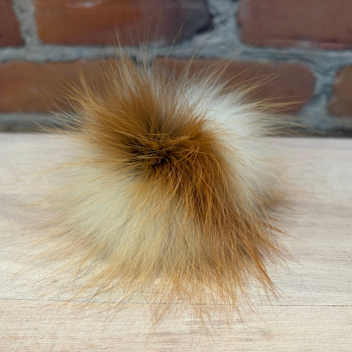 A fluffy fox fur pom-pom ball with a mix of golden brown, cream, and white tones, resting on a light wood surface with a brick wall background.
