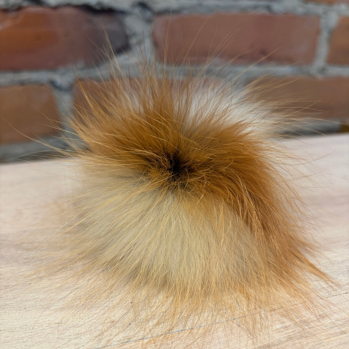 A fluffy fox fur pom-pom accessory with a golden brown top and cream-colored lower fibers, resting on a light wood surface with a blurred brick wall background.