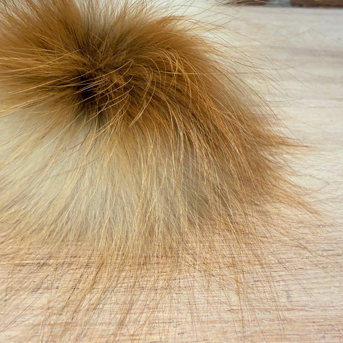 A close-up of a fluffy fox-fur pom-pom accessory with golden brown and cream tones, resting on a light wood surface with a brick wall in the background.