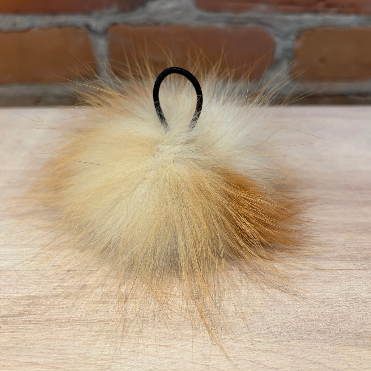 A fluffy fox fur pom-pom keychain or bag charm in beige and light brown tones, featuring a black elastic loop at the top, resting on a light wood surface with a blurred brick wall background.
