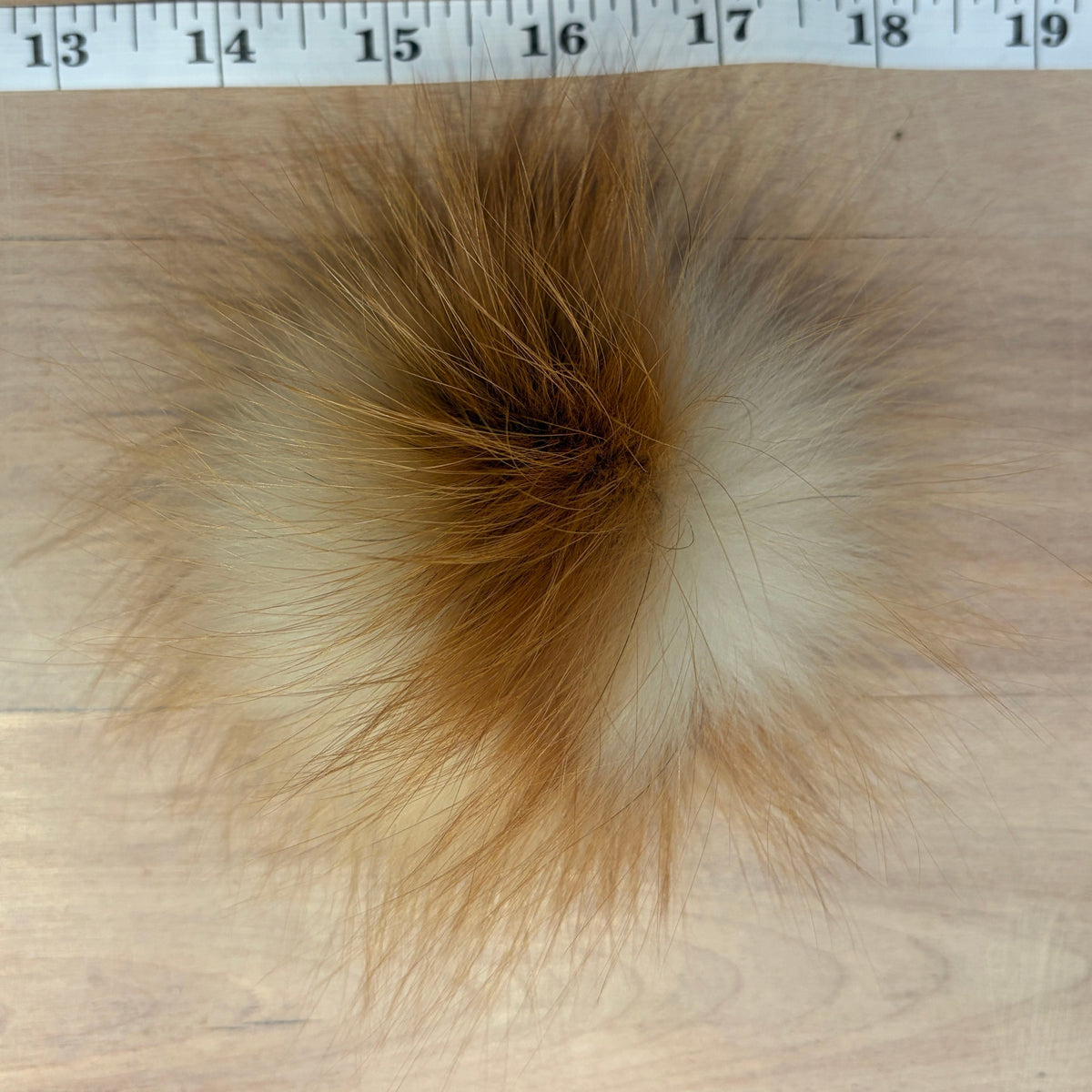 A fluffy fox -fur pom-pom with a natural fox-like coloration of brown, beige, and cream, shown on a light wood surface with a measuring tape ruler at the top for scale.