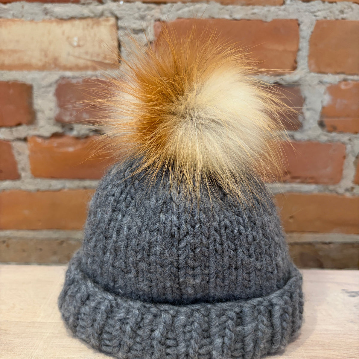 A small dark gray chunky knit beanie with a small fluffy pom-pom in shades of tan, cream, and brown, displayed on a light wood surface against a brick wall background.