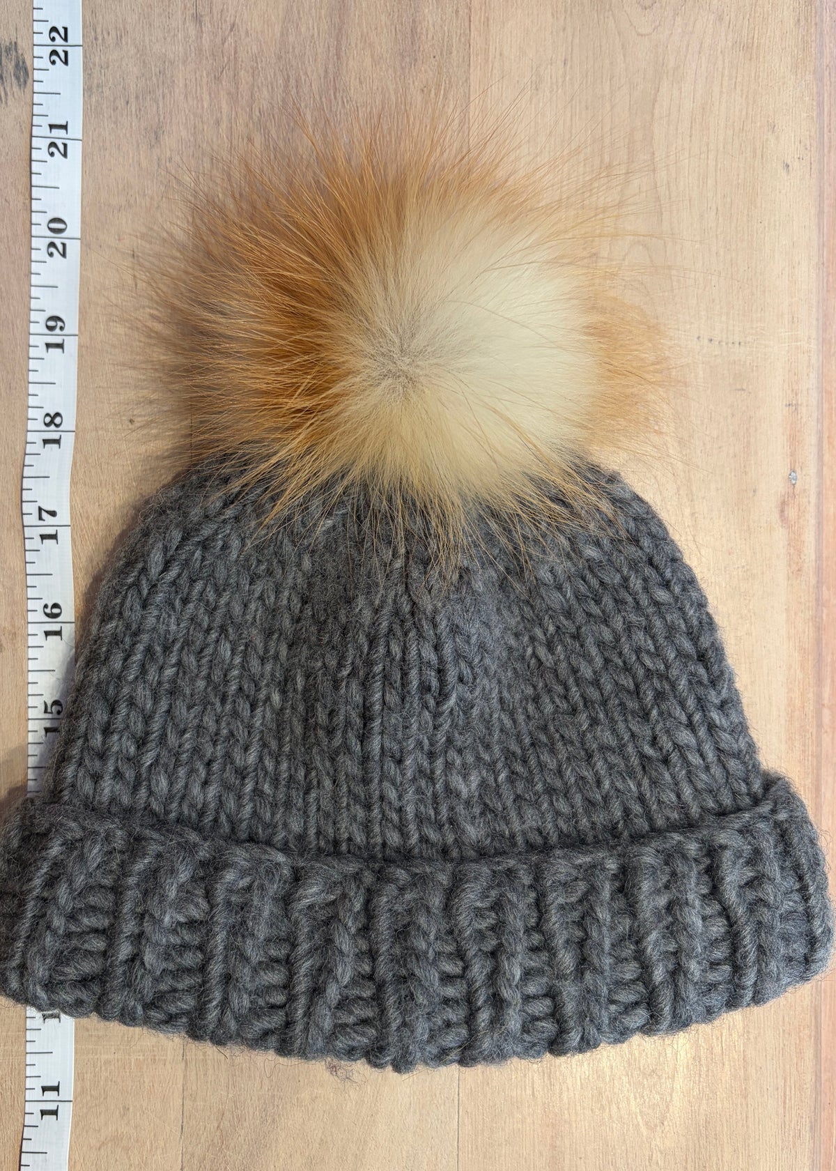 A small gray chunky knit beanie with a small fluffy pom-pom in tan and cream, laid flat on a light wood surface next to a measuring tape.