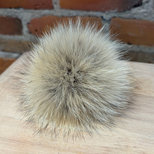 A fluffy real coyote fur pom-pom ball in shades of beige, cream, and gray, resting on a light wood surface with a brick wall background.