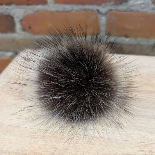 A round, fluffy raccoon-fur pom-pom accessory in dark brown and silver-gray tones, resting on a light wood surface with a blurred brick wall background.