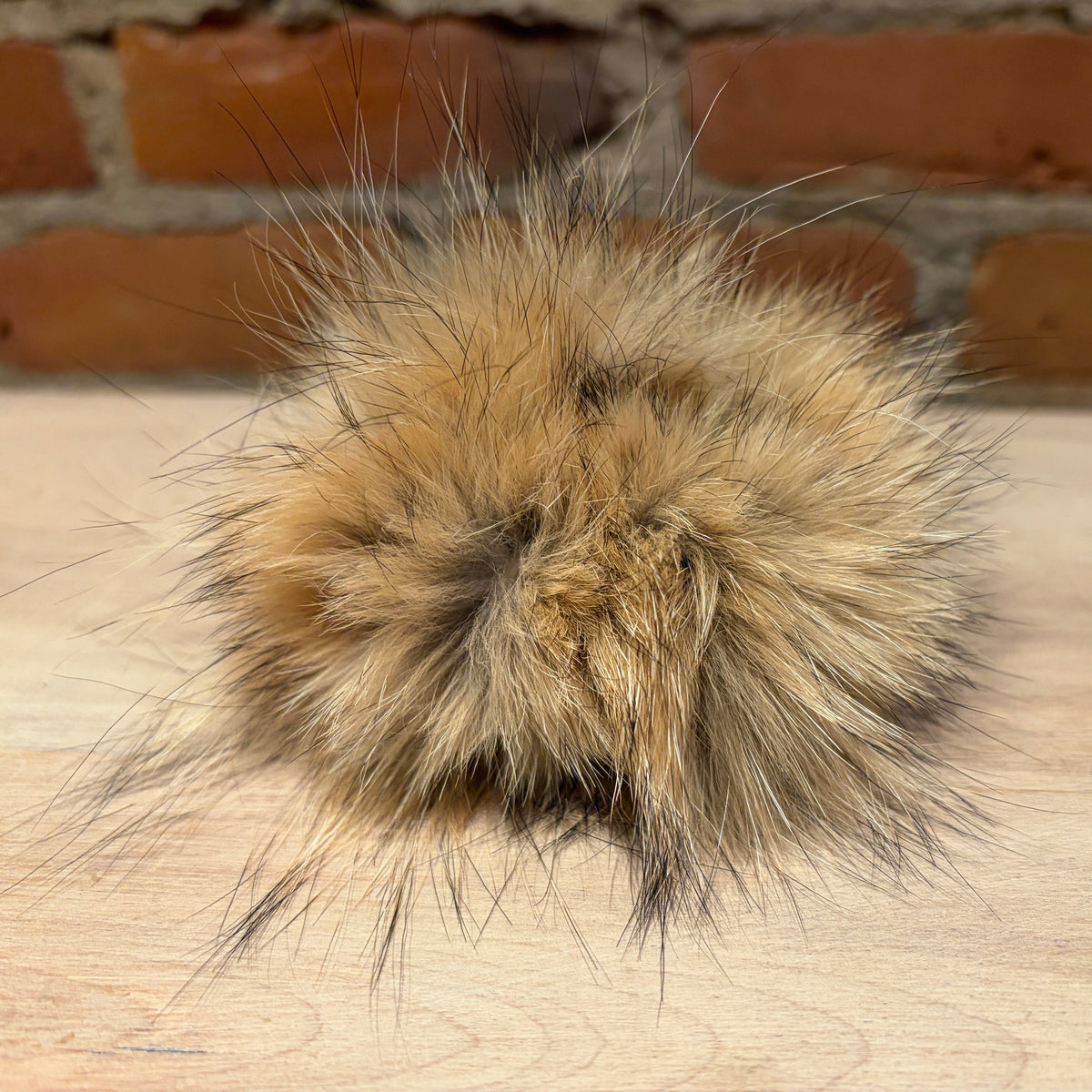 A fluffy coyote fur scrunchie accessory in warm tan and brown tones, photographed on a light wood surface with a brick wall in the background.