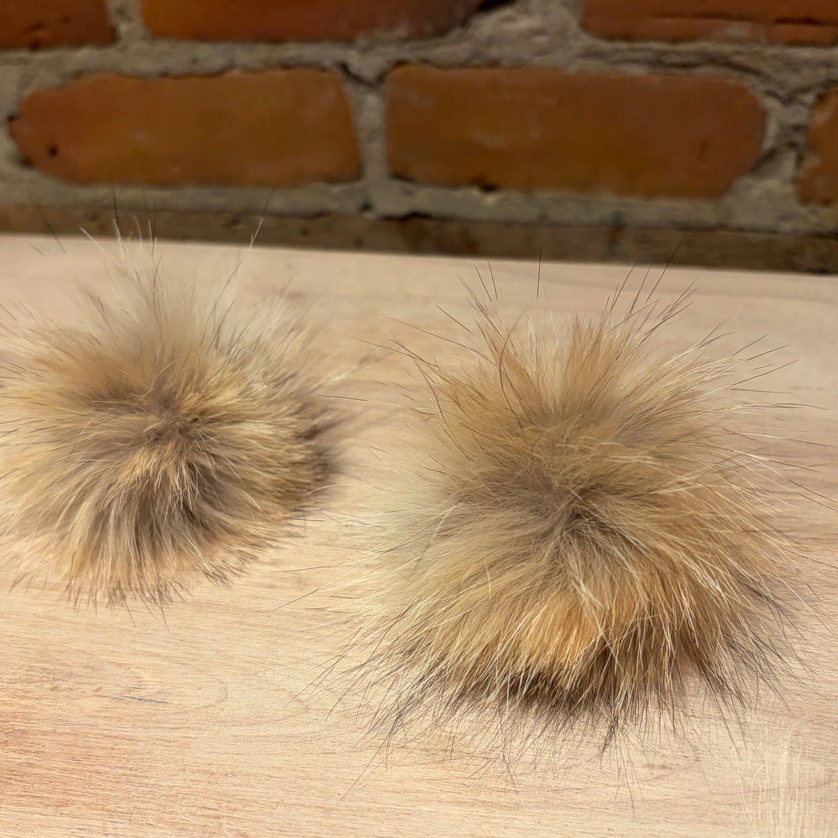 Two fluffy beige faux fur pom-poms displayed on a light wood surface with a brick wall background.