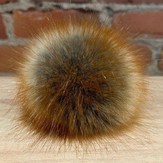 A round faux red fox faux fur pom on a wooden surface, with a blurred background.