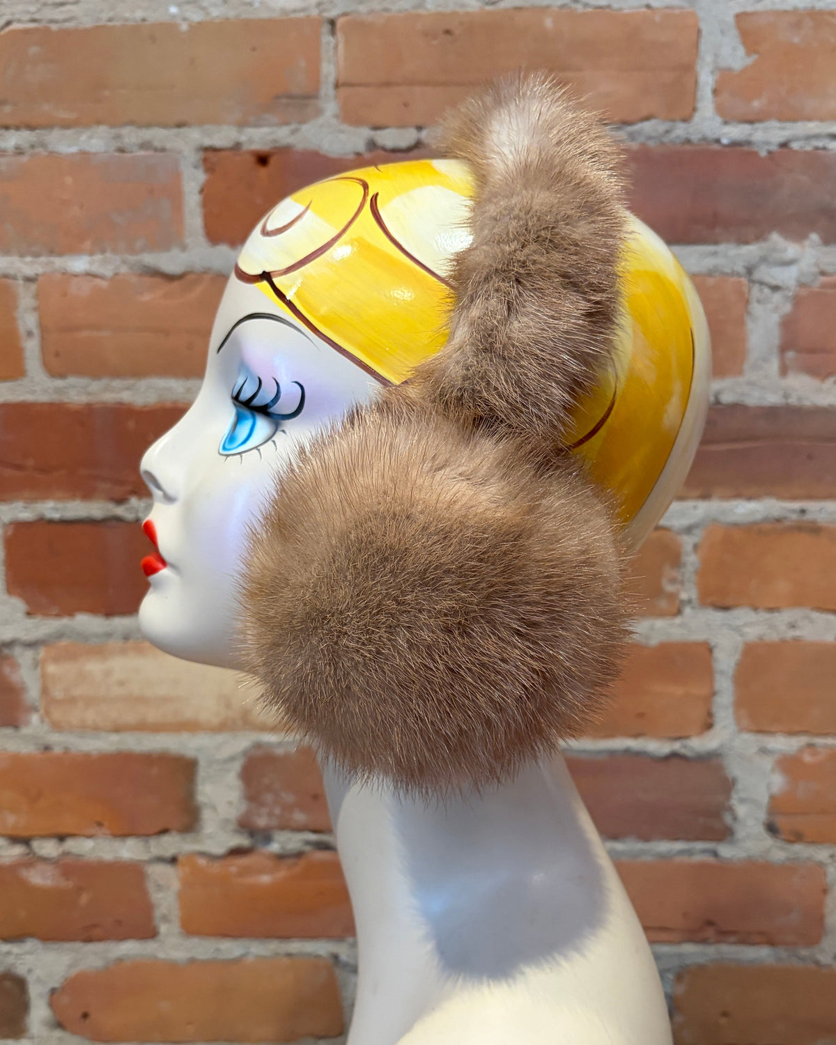 A pair of honey beige min fur earmuffs with a matching fluffy hair accessory, displayed on a vintage-style female mannequin head with a yellow and white pattern, set against a red brick wall background.
