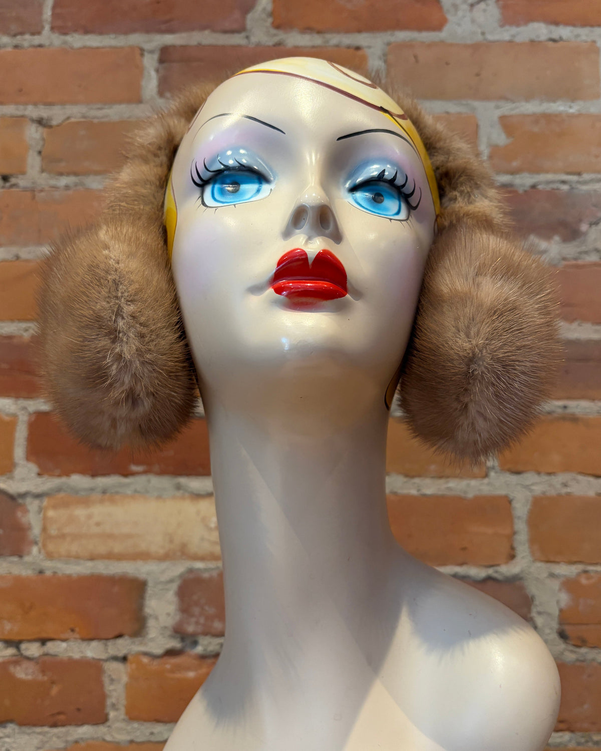 A mannequin bust wearing a pair of honey beige wraparound headband earmuffs, posed against a red-brown brick wall.