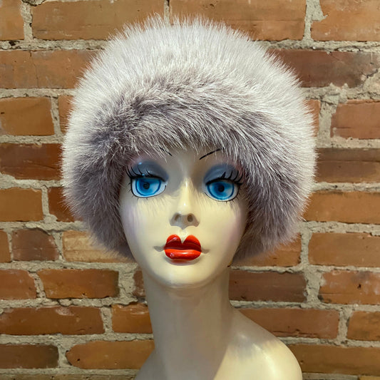 A fluffy light gray faux fur hat displayed on a mannequin head with bright blue eyes and red lips, set against a brick wall background.