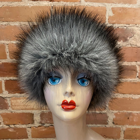 A fluffy gray faux fur headband with a black top and bottom, displayed on a mannequin head with bright blue eyes and red lips, set against a brick wall background.