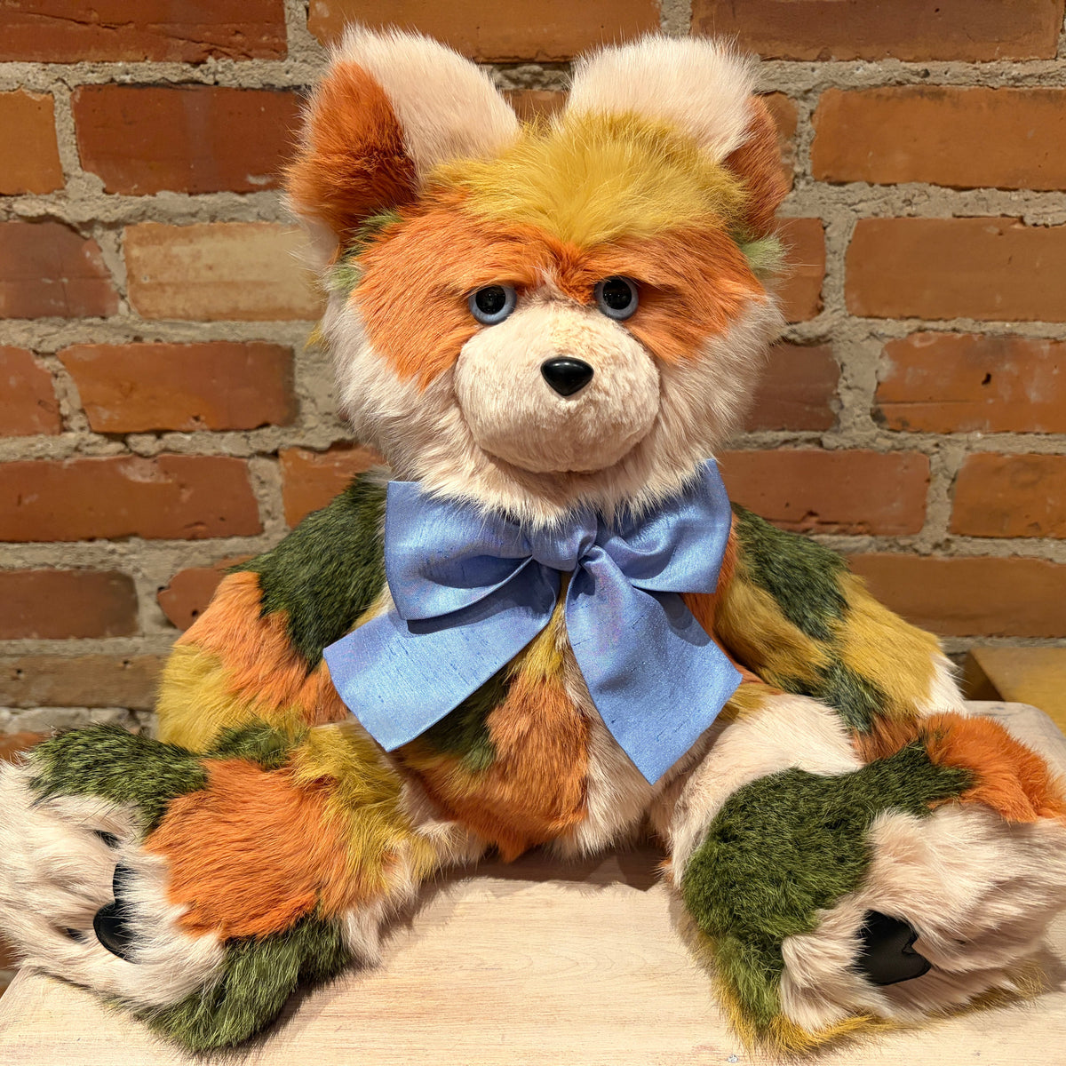A plush stuffed animal shaped like a fox or dog with orange, cream, and green patchwork fur, a light blue bow around the neck, and black eyes, posed against a brick wall background.