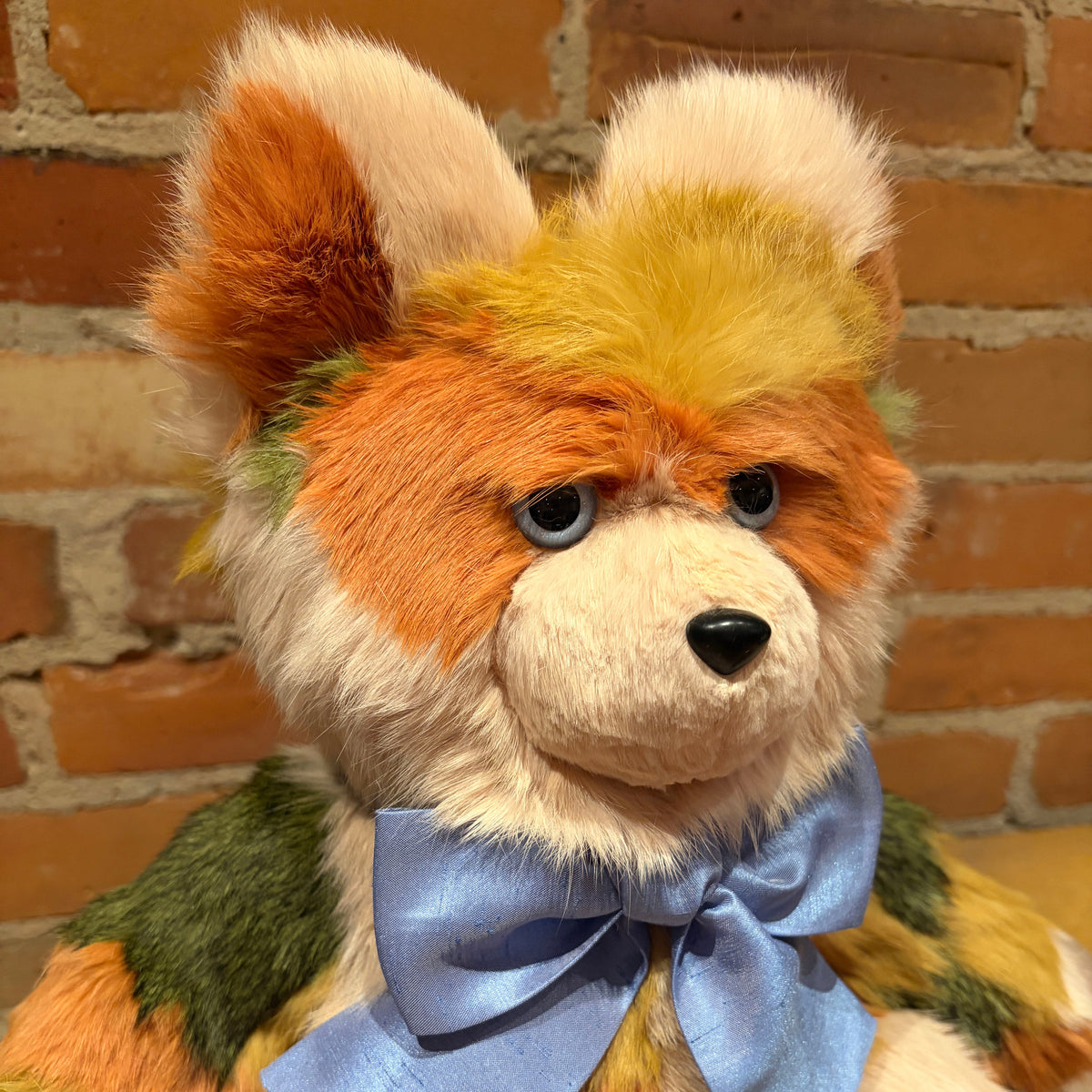 A close-up of a handmade, multi-colored plush fox toy with orange, yellow, green, and cream fur, wearing a light blue satin bow tie, set against a brick wall background.