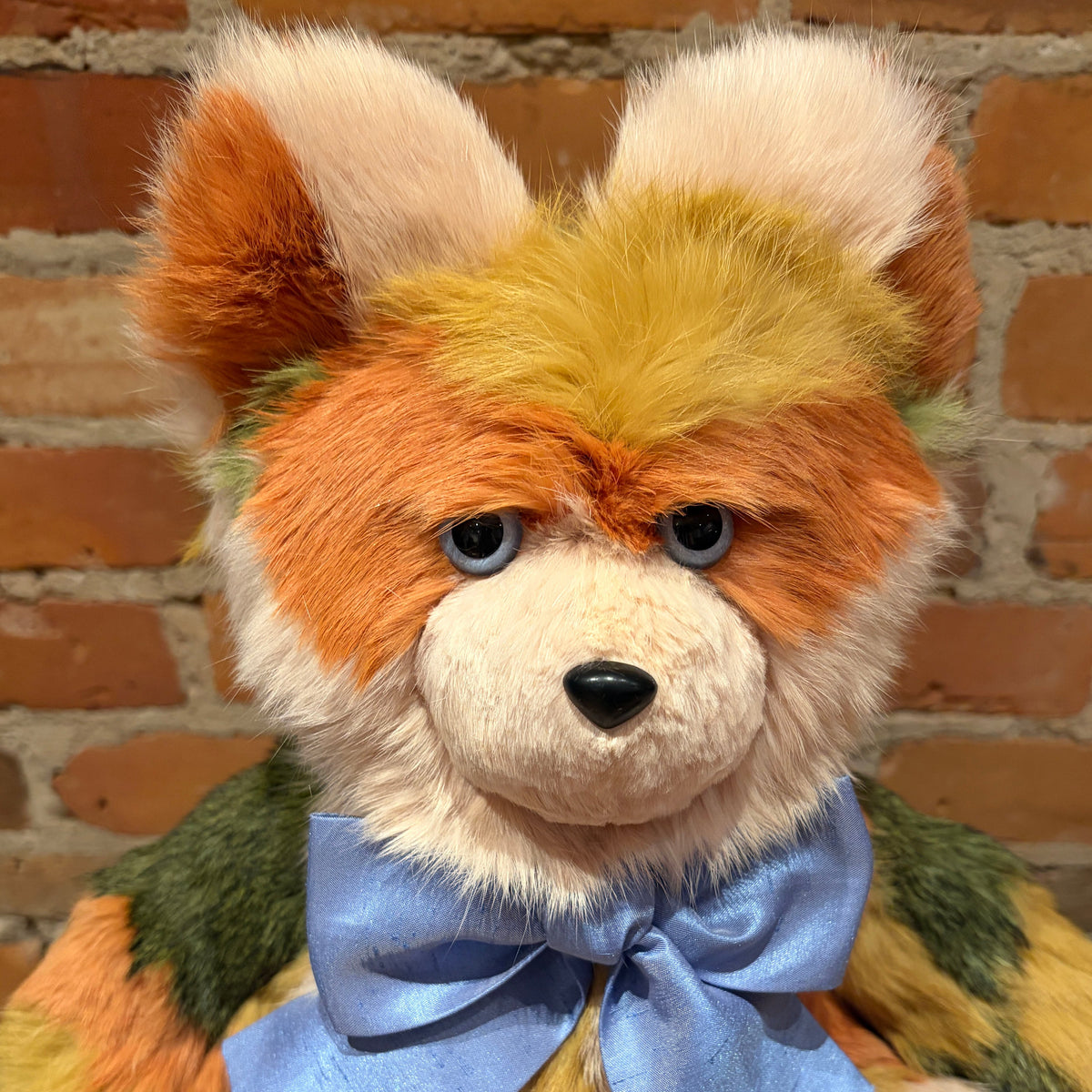 A plush teddy bear with orange, cream, and yellow fur, wearing a light blue satin bow tie, set against a brick wall background.