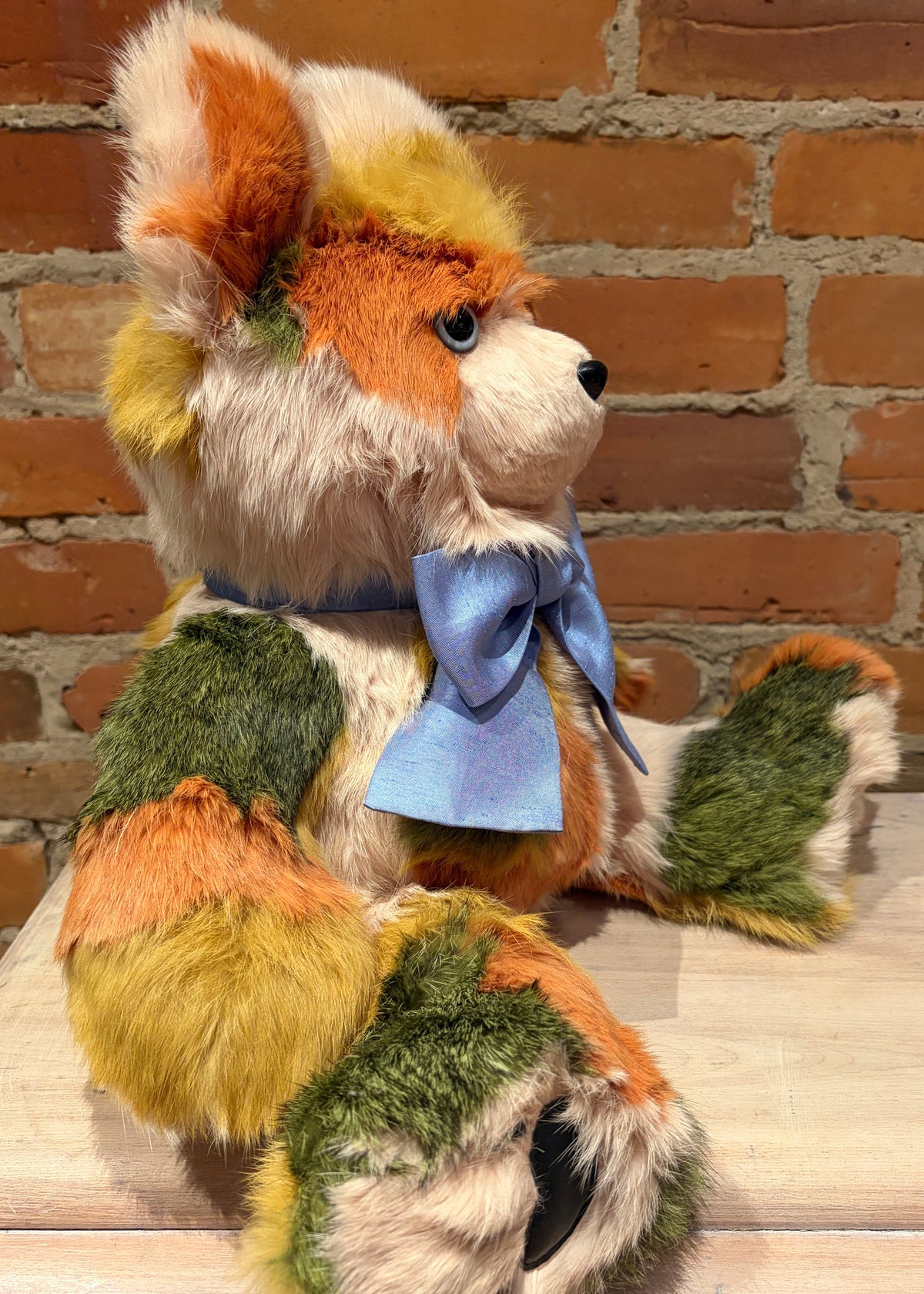 A handmade patchwork plush teddy bear with orange, green, yellow, and white fur, wearing a light blue bow, sitting in front of a brick wall.