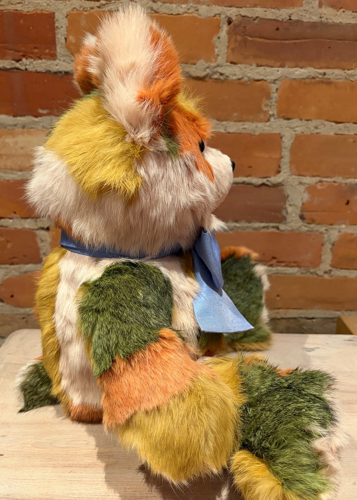 A handmade patchwork teddy bear  shaped like a fox or cat, sitting on a light wooden surface against a rustic red brick wall. The plush features multiple colored fur patches including green, yellow, orange, and cream, with a light blue ribbon tied around its neck.