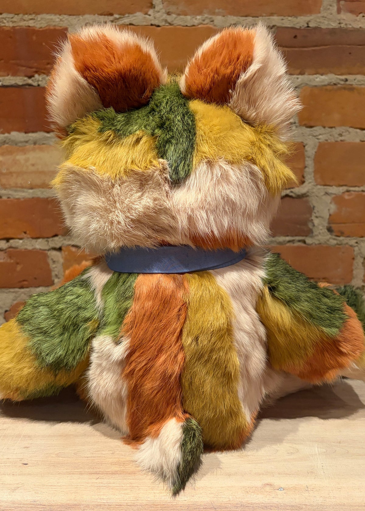 A handmade, multi-colored teddy bear shaped like a fox or cat, made from patchwork fur in orange, green, yellow, and cream, sitting in front of a brick wall.