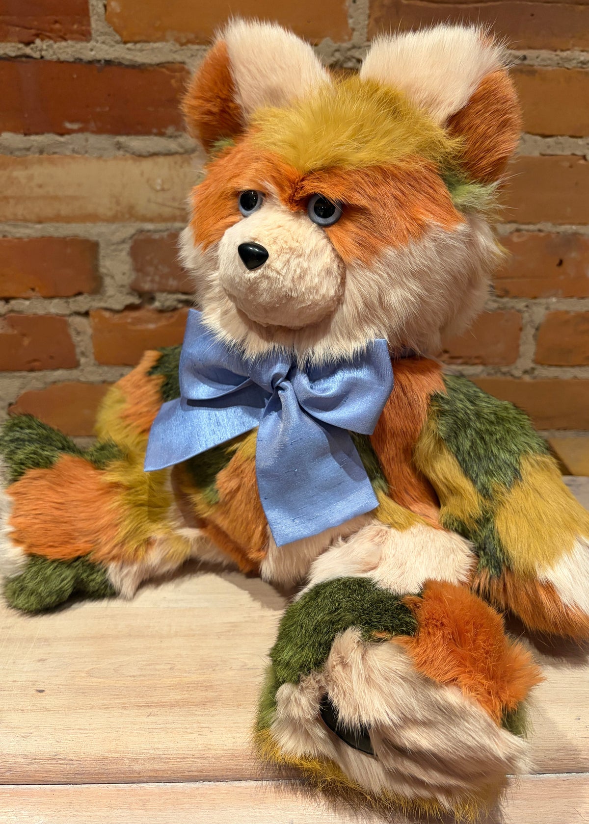 A plush handmade teddy bear with orange, cream, and green patchwork fur, wearing a light blue bow tie, posed on a wooden surface against a brick wall background.