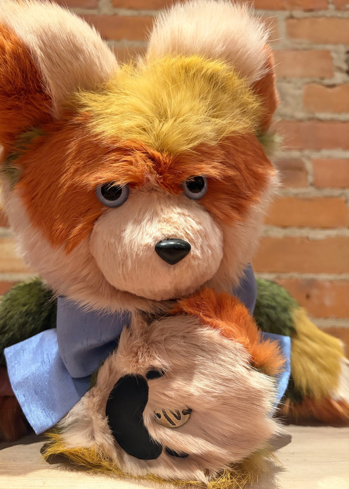A close-up of a plush stuffed animal with orange, cream, and yellow fur, wearing a blue bow tie, posed in front of a brick wall background.