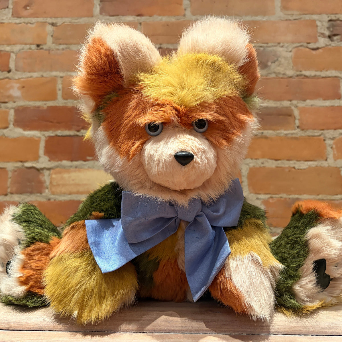 A handmade plush teddy bear with multi-colored fur in shades of orange, yellow, green, and cream, wearing a blue bow tie, posed against a rustic brick wall background.
