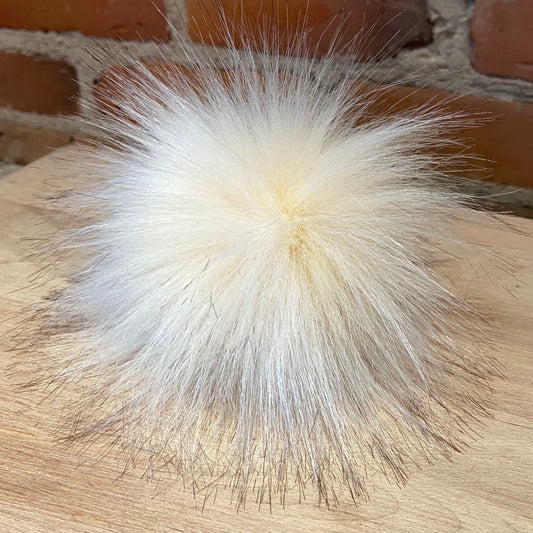 A fluffy, voluminous pom-pom in a warm cream or off-white shade, resting on a light wood surface with a rustic red brick wall in the background.