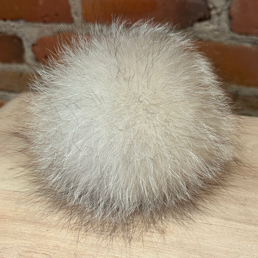 A fluffy pom-pom ball in light cream or off-white fox fur, resting on a light wood surface with a blurred brick wall background.