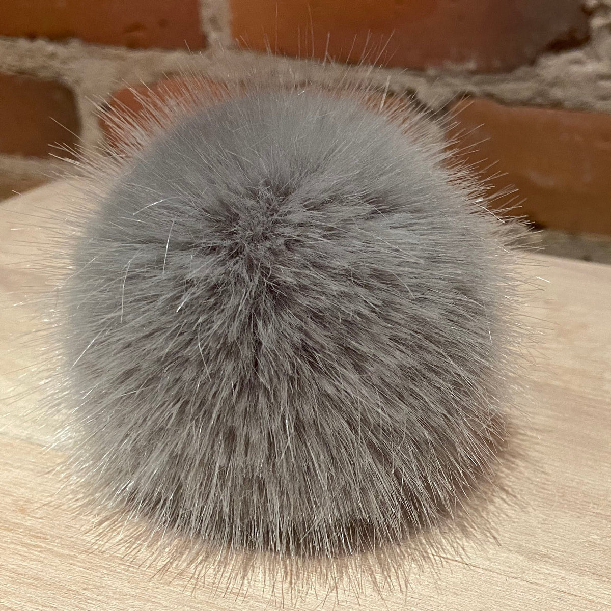 A fluffy gray faux fur ball accessory resting on a light wood surface with a brick wall background.