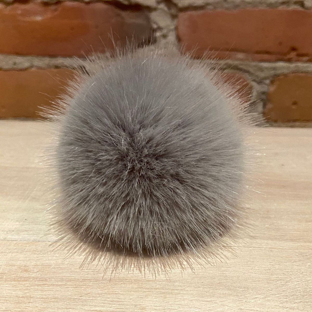 A round, fluffy gray faux fur pom-pom accessory resting on a light wood surface with a brick wall background.