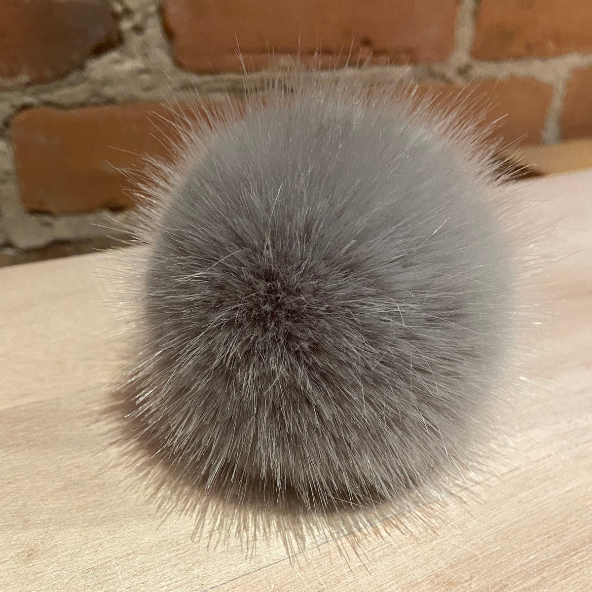 A fluffy gray faux fur pom-pom ball resting on a light wood surface with a brick wall background.