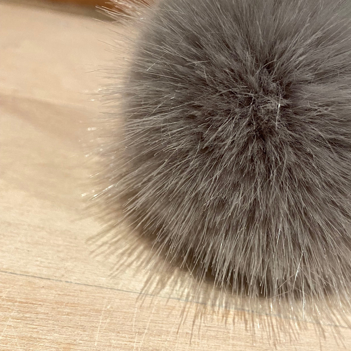 A close-up of a fluffy gray faux fur pom-pom resting on a light wood surface with a blurred brick wall in the background.
