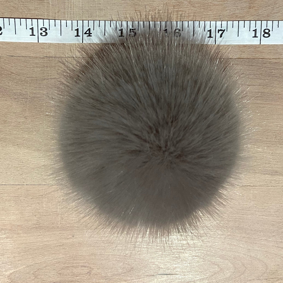 A round gray faux fur pom-pom ball shown on a light wood surface with a white measuring tape ruler at the top for scale.