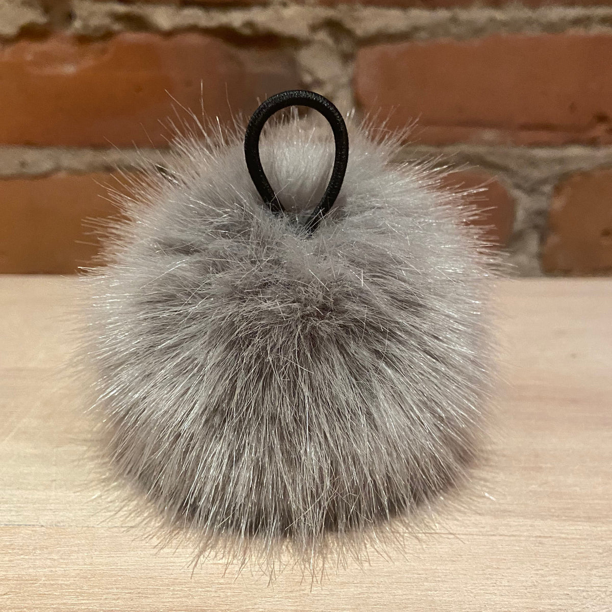 A fluffy gray faux fur hat pom with a black hanging loop, resting on a light wood surface with a brick wall background.