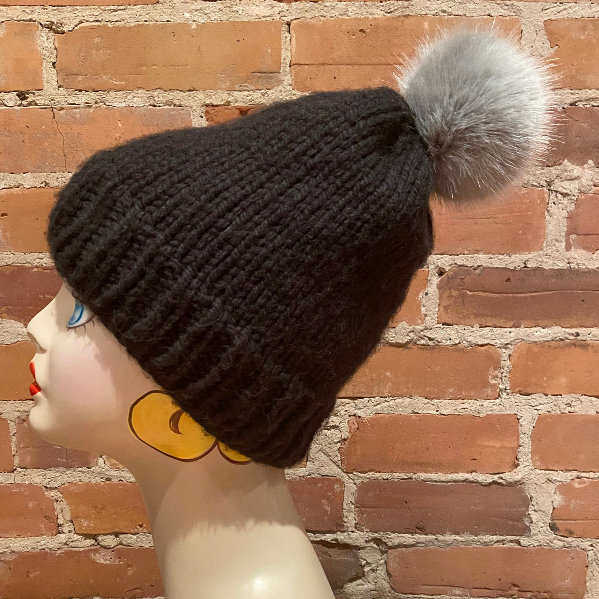 A black ribbed knit beanie with a fluffy light gray pom-pom, displayed on a mannequin head against a red brick wall background.