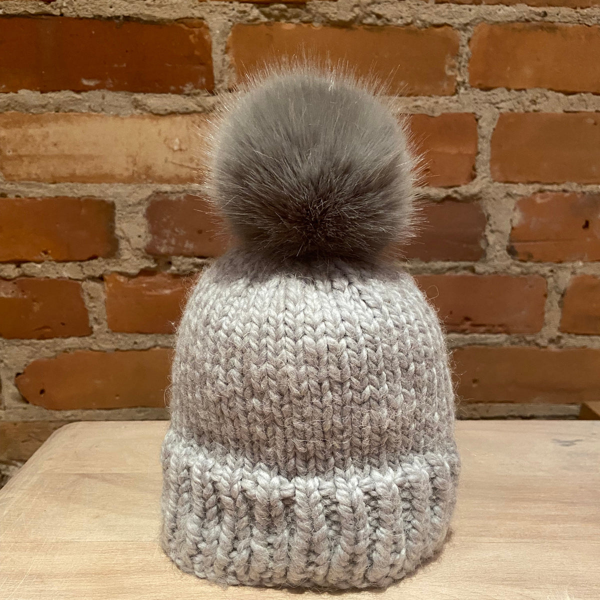 A small light gray knit beanie hat with a small fluffy taupe-gray pom-pom, displayed on a wooden surface against a brick wall background.