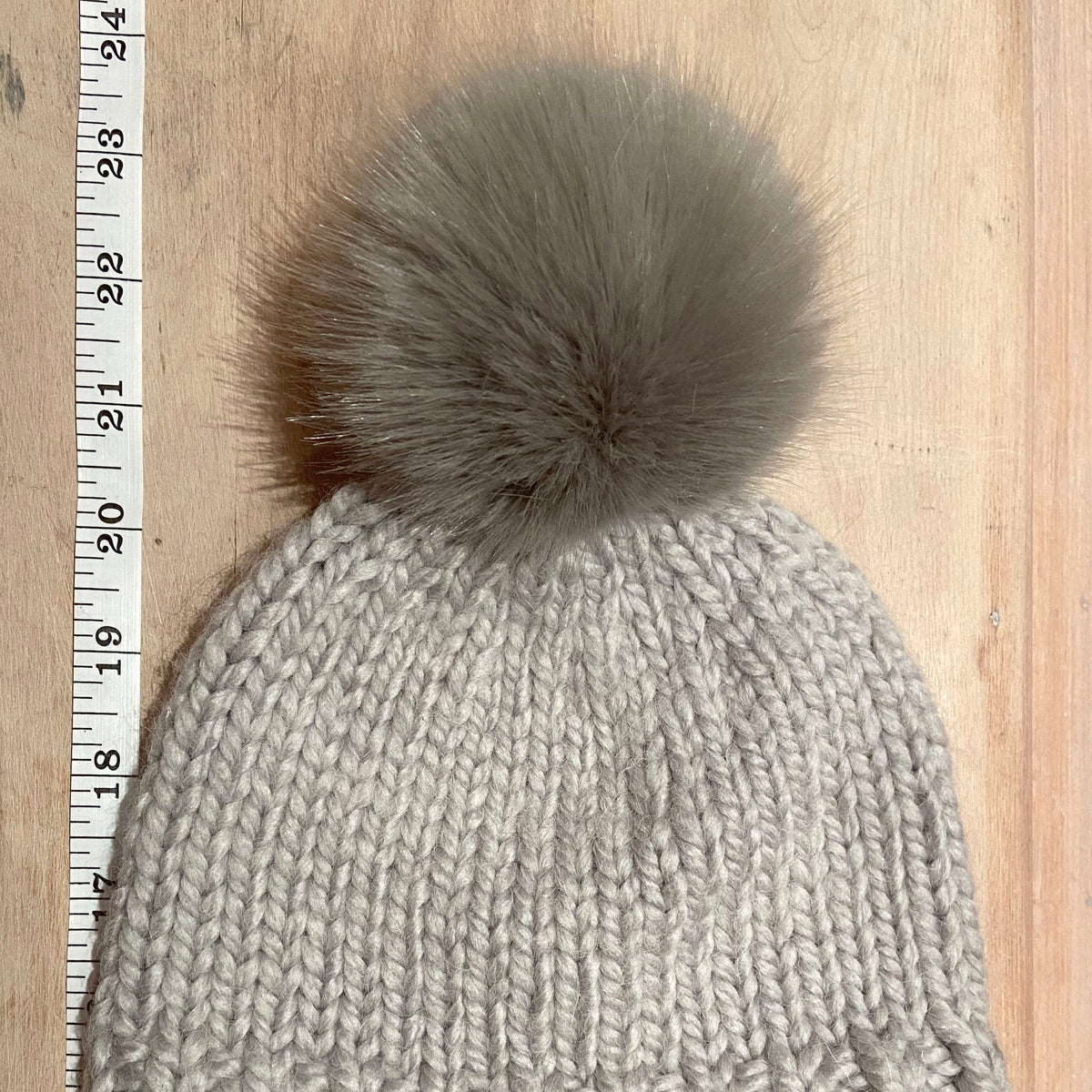 A small light gray chunky knit beanie with a small taupe-gray faux fur pom-pom, laid flat on a light wood background next to a measuring ruler.