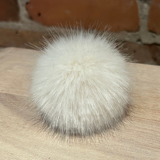 A fluffy white faux fur pom-pom ball resting on a light wood surface with a rustic brick wall background.