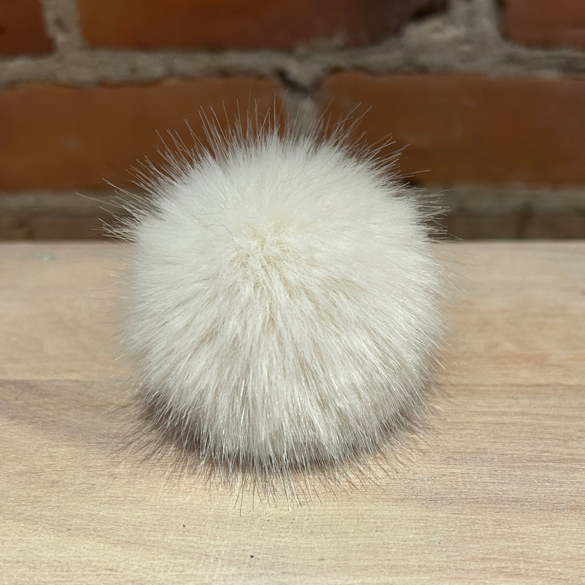 A fluffy white faux fur pom-pom ball resting on a light wood surface with a blurred brick wall background.