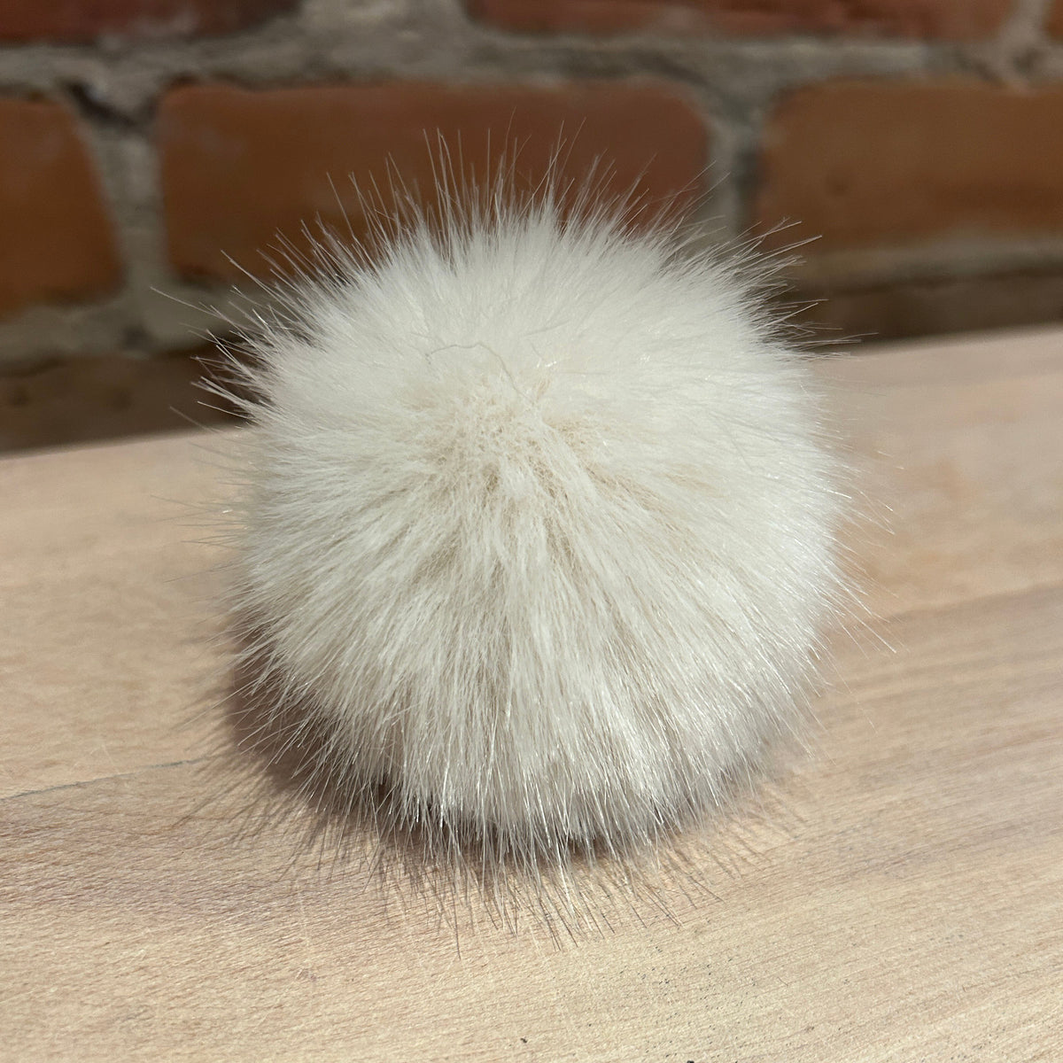 A fluffy white faux fur pom-pom accessory resting on a light wood surface with a blurred brick wall background.