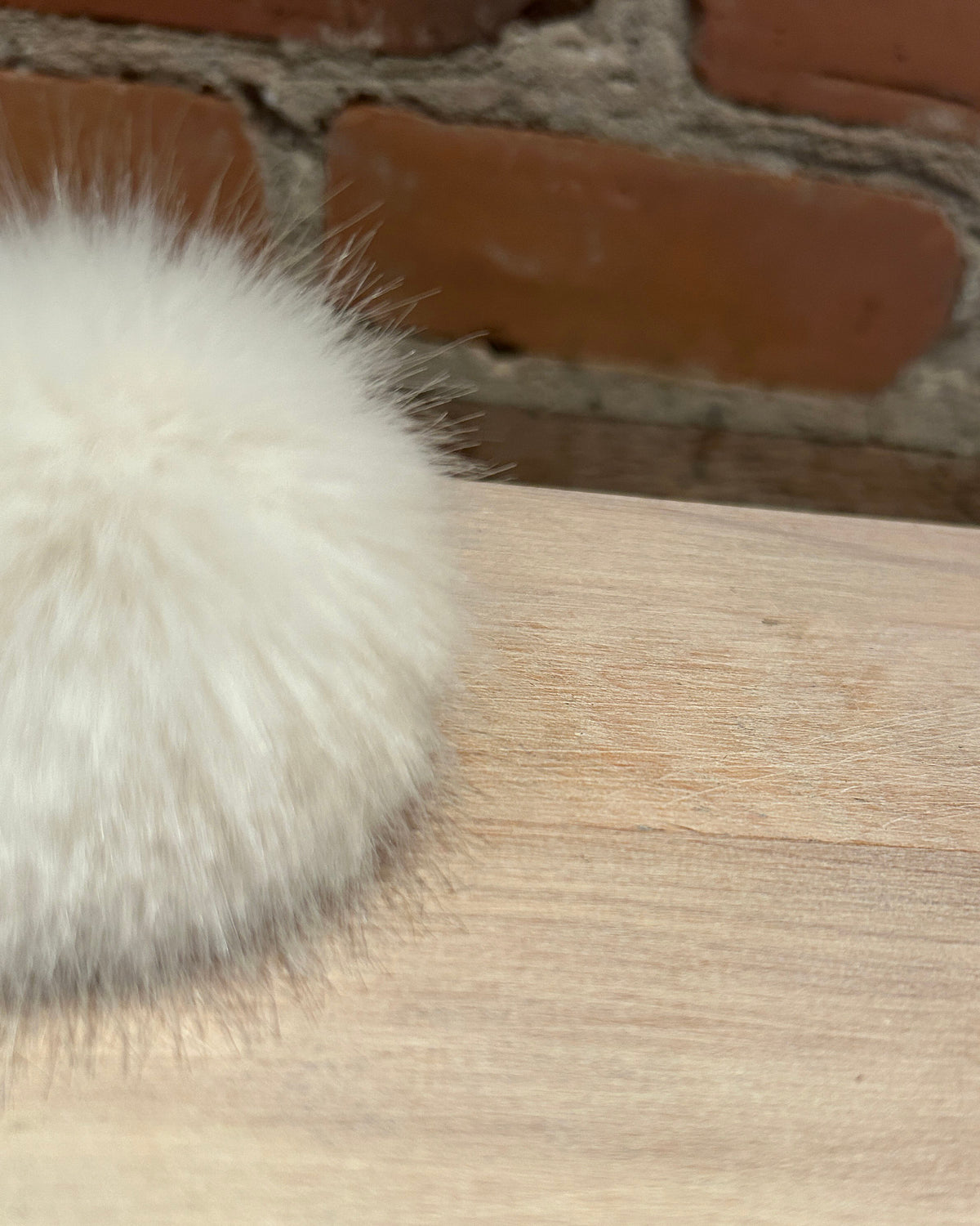 A close-up of a fluffy white faux fur pom-pom resting on a light wood surface with a brick wall in the background.