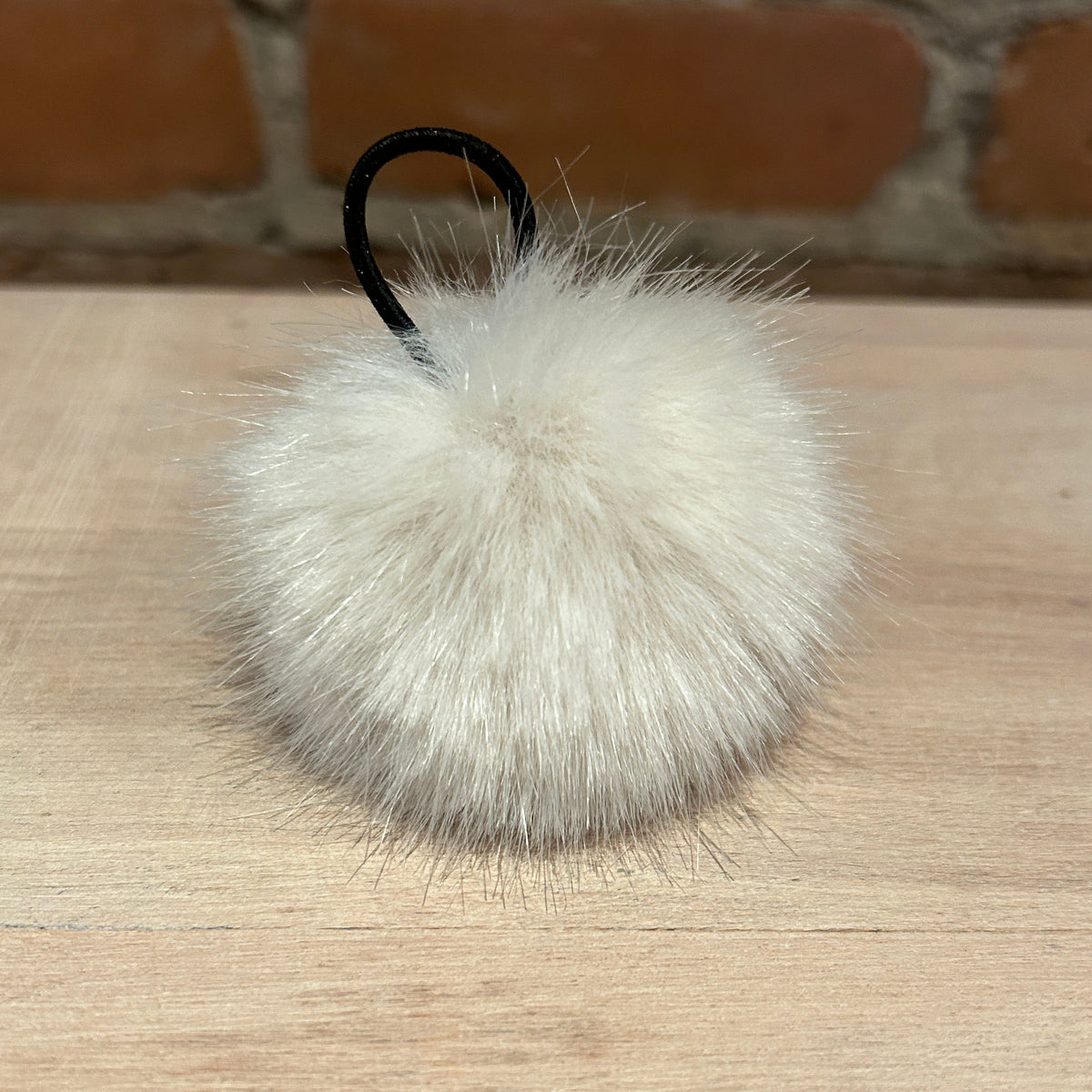 A fluffy white faux fur pom-pom keychain with a black elastic loop, resting on a light wood surface with a blurred brick wall background.