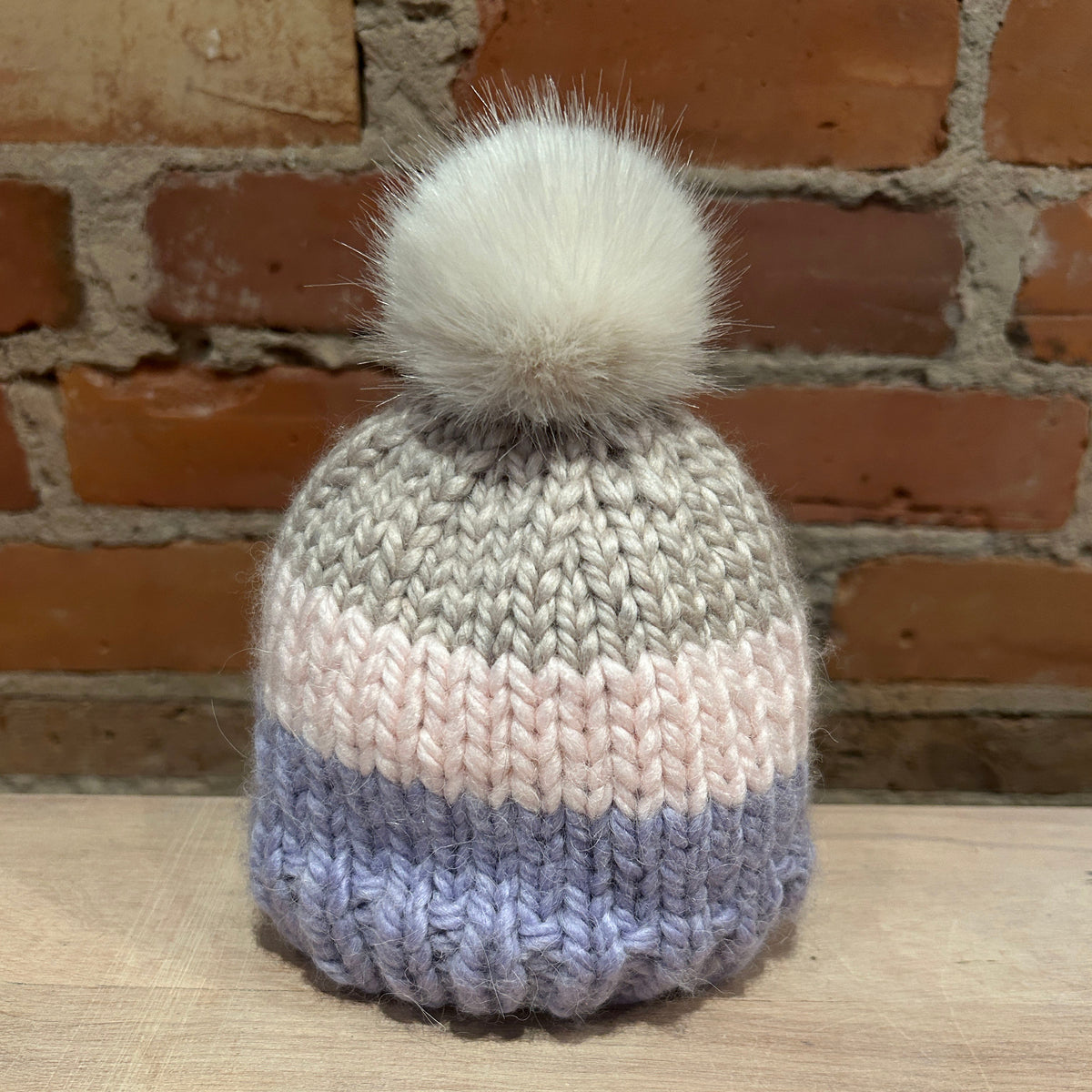 A small hand-knit beanie hat with a small fluffy pom-pom, featuring horizontal stripes of gray, pale pink, and lavender blue, displayed on a wooden surface against a brick wall background.