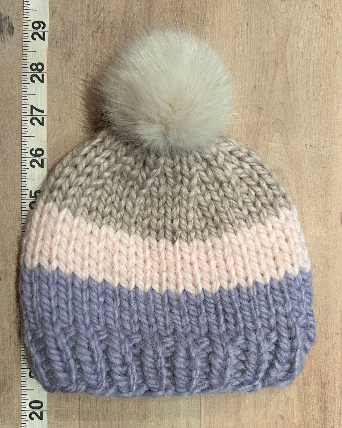 A hand-knit beanie hat with three horizontal color bands: taupe, pale pink, and dusty purple, topped with a fluffy cream pom-pom. The beanie is laid flat on a light wood surface next to a measuring tape ruler.