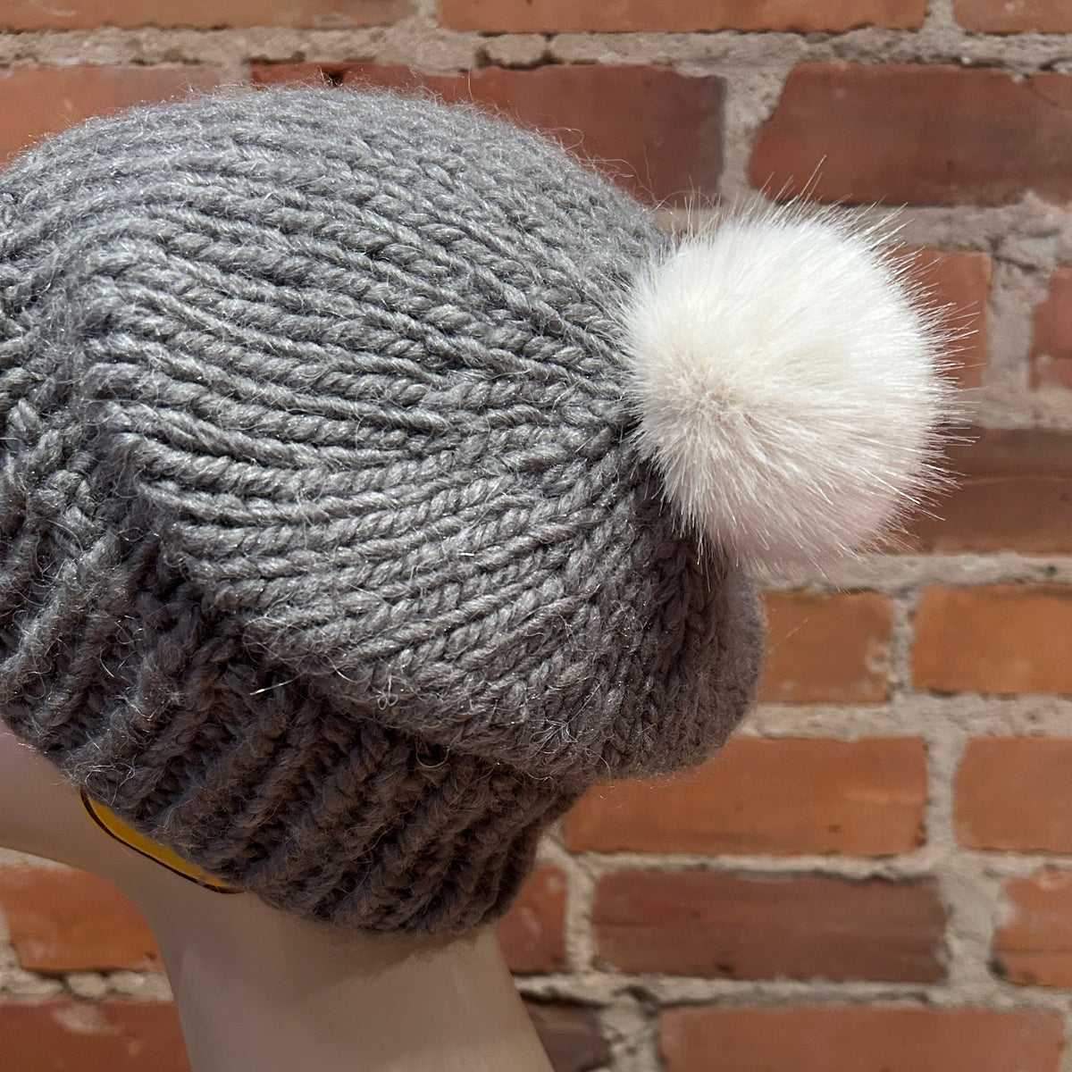 A gray chunky-knit beanie hat with a fluffy white pom-pom, displayed on a mannequin head against a red brick wall background.